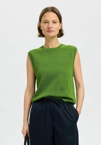 Grünes ärmelloses Oberteil mit Rundhalsausschnitt, strukturiertem Stoff und lässiger Passform, kombiniert mit navyblauen Hosen. Das Modell hält eine kleine schwarze Tasche.