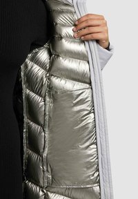 Vest s náplňou so lesklým strieborným vnútorným materiálom a prequiltnou textúrou, orámované mäkkou bielou vonkajšou vrstvou a elegantnou zipsovou kapsou.