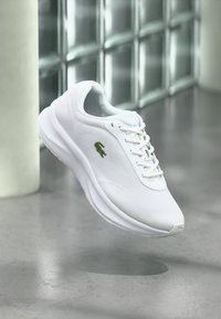 Άσπρο αθλητικό παπούτσι Lacoste με πράσινο λογότυπο κροκόδειλου, να αιωρείται πάνω από μια γυαλισμένη τσιμεντένια επιφάνεια κοντά σε παράθυρα από γυάλινα τούβλα.