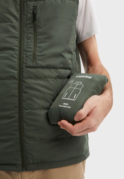 Osoba v olivově zelené zateplené vestě, která před bílým pozadím drží kompaktní tašku označenou "Tropicfeel NS40 Reversible Vest".