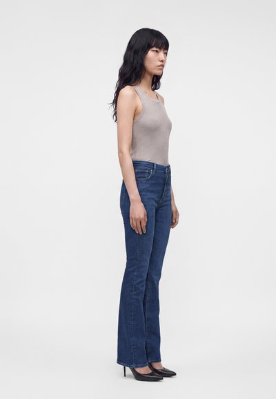 Femme aux longs cheveux noirs portant un haut beige sans manches, un jean bleu taille haute et des talons noirs, debout de profil.