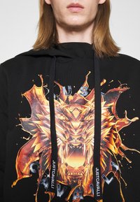 Schwarzer Kapuzenpullover mit einem farbenfrohen Grafikdesign eines Löwenkopfes, ergänzt durch orange und gelbe Spritzeffekte, metallische Buchstaben und schwarze Kordeln.