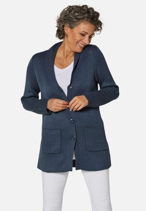 Marineblauer Cardigan aus strukturiertem Stoff, mit einem Schalkragen, Knopfverschluss und zwei Fronttaschen, über einem weißen Hemd getragen.