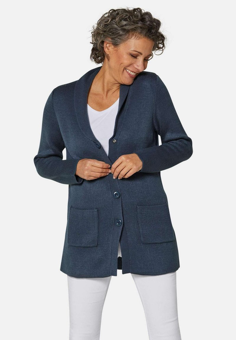 Cardigan bleu marine en tissu texturé, doté d'un col châle, d'une fermeture à boutons et de deux poches avant, porté sur une chemise blanche.