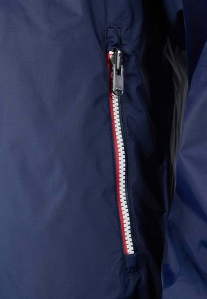 Chaqueta azul marino con cremallera impermeable diagonal y pestaña blanca y roja. Textura de tela lisa con un sutil patrón de cuadricula.