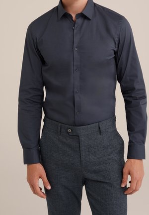 Mann trägt ein eng anliegendes, dunkelblaues Hemd mit Knöpfen, in graue karierte Anzughose gesteckt, steht vor einem neutralen Hintergrund.