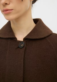 Cappotto di lana marrone con colletto arrotondato, dotato di chiusura a singolo bottone e un tessuto morbido e testurizzato. Nessun motivo o accento visibile.