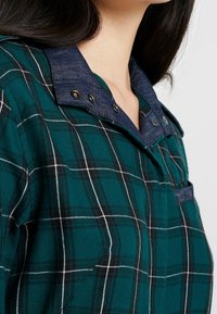 Grünes Karoshemd mit weißen und schwarzen Linien, versehen mit einem Denim-Kragen und einer Tasche. Knopfdetails betonen den Kragen. Weiches, strukturiertes Gewebe.