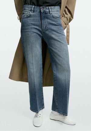 Jean bleu droit à usure moyenne, associé à des chaussures blanches à lacets, un trench beige et un haut noir avec les mains dans les poches.
