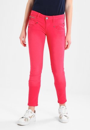 Mujer con pantalones ajustados de color rosa brillante y zapatillas blancas con estampado animal, de pie contra un fondo claro y liso.