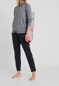 Graues Langarmoberteil mit schützendem Kragen, kombiniert mit schwarzen Jogginghosen mit Kordelzug. Eine rosa Yogamatte ist über die Schulter gehängt.