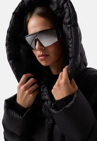 Sort pufferjakke med høj krave og hætte, med quiltet tekstur. Spejlede oversized solbriller fuldender looket. Hudtonen er neutral.