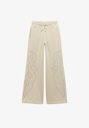 Pantalones beige de pierna ancha con cintura ajustable con cordón, bolsillos laterales y patrones cosidos en forma de llamas negras en la parte delantera de las piernas.