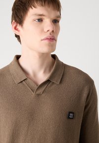 Brązowa, teksturowana koszulka polo z dekoltem w serek i krótkimi rękawami, z czarną gumową naszywką logo na lewej części klatki piersiowej.