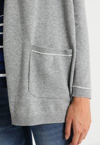 Grå stickad cardigan med framficka, bar över en randig skjorta, tillsammans med blå jeans, som visar detaljer på höger hand och handled.