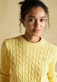 Jauna moteris su garbanotais plaukais, dėvinti auksinius auskarus-žiedus ir geltoną mezginį su raštinėmis, turi mažą mėlyną siuvinėtą detalę.