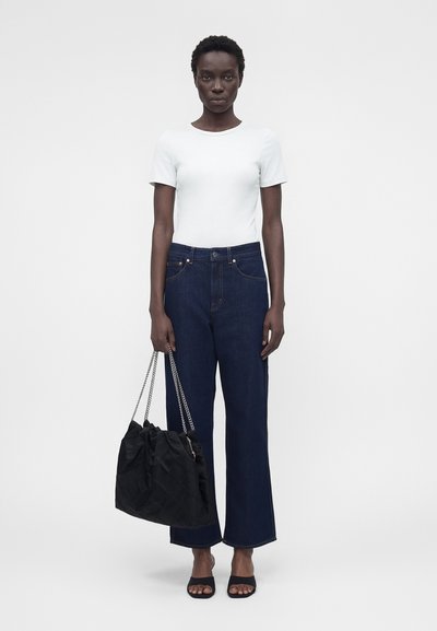 Γυναίκα όρθια, φοράει λευκή κορυφή με κοντά μανίκια, σκούρα μπλε παντελόνι με φαρδιά πόδια, μαύρα ανοιχτά τακούνια, κρατά μεγάλη μαύρη τσάντα με αλυσίδες.