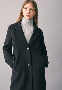 Cappotto di lana nera con colletto a revers, chiusura a singolo bottone e tasche laterali, sovrapposto a un dolcevita grigio chiaro a coste.