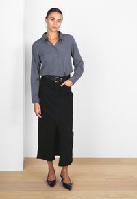 Chemise grise à boutons, jupe noire avec une fente sur le devant, chaussures à talons hauts noires, collier en chaîne dorée et boucles d'oreilles créoles. Arrière-plan uni.