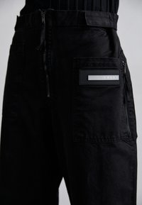 Pantalones cargo negros con un bolsillo con cremallera y una etiqueta rectangular texturizada que contiene la palabra "ARIES". Fabricados con tejido duradero, diseño de pierna recta.