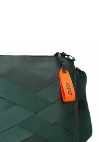 Grüne Stofftasche mit ineinander geflochtenen Trägern, einem Reißverschluss und einem leuchtend orangefarbenen Etikett mit Branding. Die Textur ist glatt und langlebig.