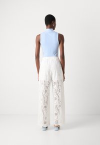 Top senza maniche di un blu chiaro abbinato a pantaloni bianchi a gamba larga con un motivo floreale ritagliato; il modello si trova in uno spazio minimalista.