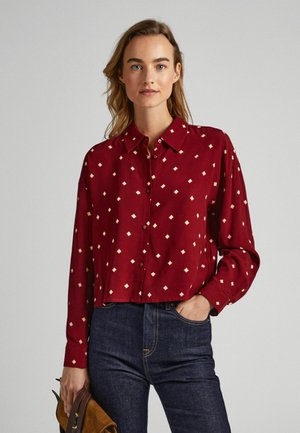 Camisa de botón color burdeos con corte relajado, con patrones de diamante blancos. Mangas largas con puños, combinada con jeans de mezclilla oscura.