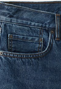 Gros plan d'une poche de jean en denim bleu avec des coutures apparentes, des rivets et une petite poche intérieure pour pièces au niveau de la ceinture.