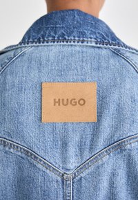 Denimjas met een blauw vervaagd patroon, voorzien van een rechthoekig bruin leren label met de tekst "HUGO" op de achterkant van de kraag.