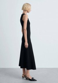 Frau mit kurzem blonden Haar, die ein ärmelloses schwarzes Midikleid und schwarze Slingback-Absätze trägt, steht im Profil vor einem schlichten Hintergrund.