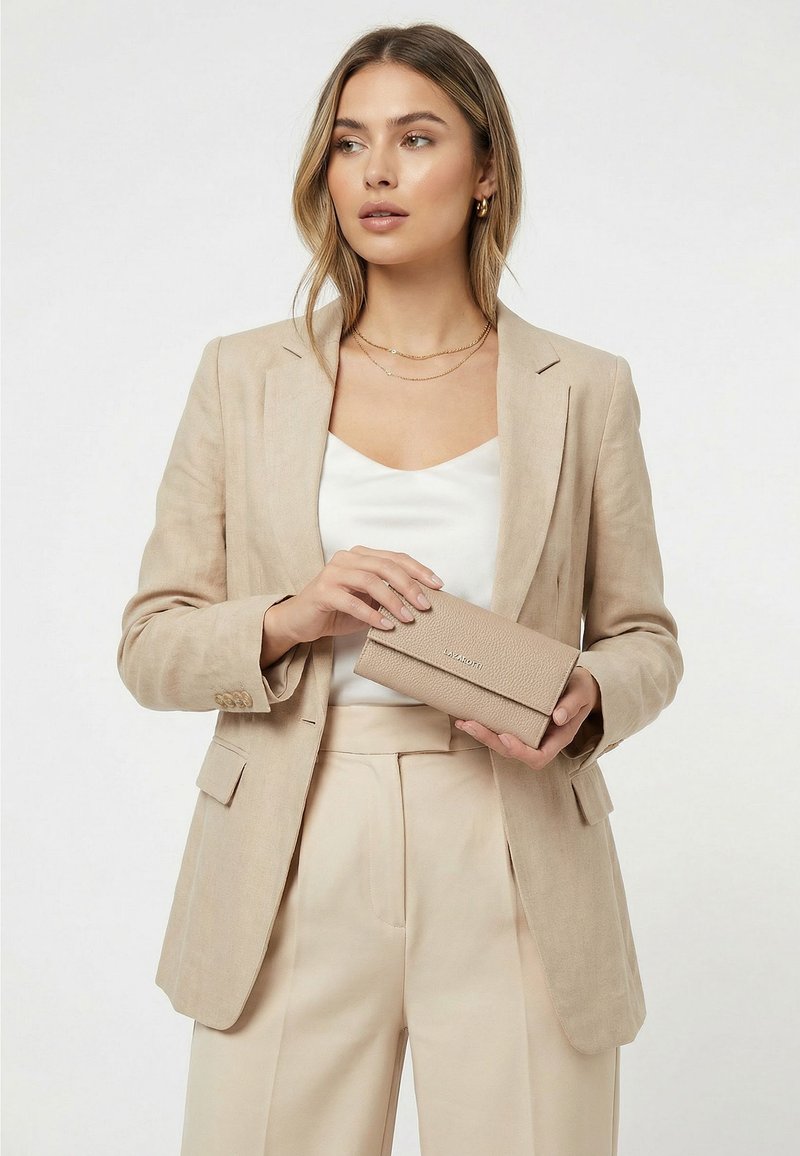 Mujer que lleva blazer y pantalones beige sostiene una cartera beige a juego mientras mira hacia un lado con un fondo liso.