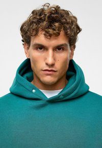 Teal hoodie met een trekkoord en drukknopdetails. De stof lijkt zacht, met een klassieke snit en een grote capuchon. Witte shirt eronder zichtbaar.
