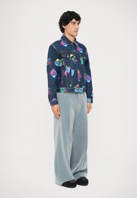 Džínová bunda s viacfarebnými grafickými potlačami, v kombinácii s nadrozmernými šedými manšestrovými nohavicami a čiernymi slip-on topánkami.