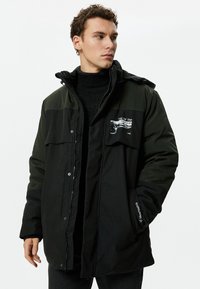 Schwarze und grüne isolierte Parkajacke mit Reisverschluss vorne, zwei Brusttaschen und besticktem Logodetail an den Ärmeln. Glatte Textur.