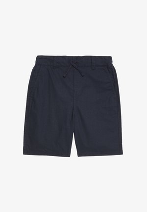 Teenager-Junge steht, trägt dunkles kurzärmeliges Hemd mit Knöpfen, passende Shorts, weiße Sportsocken und beige-weiße Turnschuhe, vor einfarbigem Hintergrund.