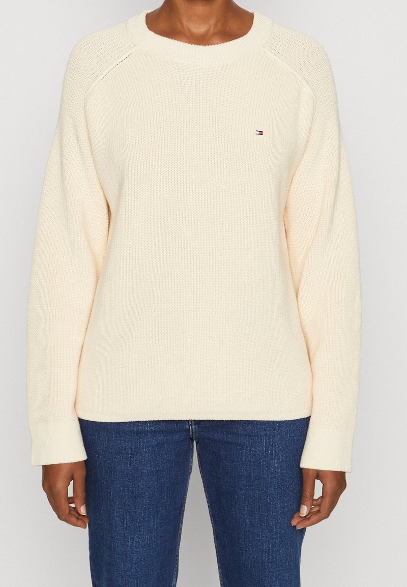 Personne portant un pull en maille crème uni à col rond et un jean bleu, debout avec les bras détendus le long du corps.