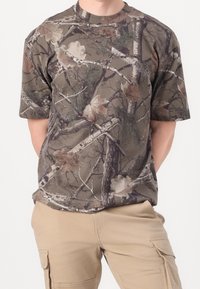 T-shirt à manches courtes en camouflage marron avec un col rond, présentant des motifs de feuilles et de branches sur un fond terrestre atténué.