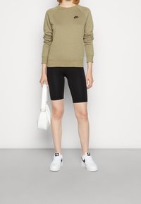 Oliivinvihreä huppari, jossa on musta Nike-logo, mustat pyöräilyshortsit, valkoiset lenkkarit ja pieni valkoinen käsilaukku. Yksinkertainen muotoilu ja rento istuvuus.