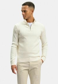 Krämfärgad kabelstickad tröja med kvartdragkedja, ribbade ärmslut och nederkant. Bärs över en vit t-shirt, i kombination med ljusbeige byxor.