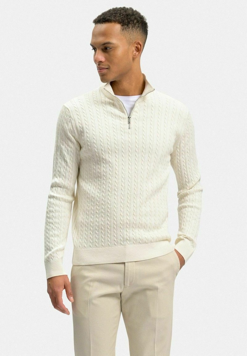 Krämfärgad kabelstickad tröja med kvartdragkedja, ribbade ärmslut och nederkant. Bärs över en vit t-shirt, i kombination med ljusbeige byxor.