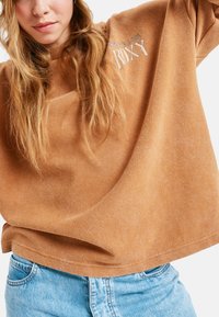 Luźna, waflowa bluzka z długim rękawem w kolorze terakoty z białym logo "Roxy". Połączona z jasnoniebieskimi szortami dżinsowymi, prezentując swobodny styl.