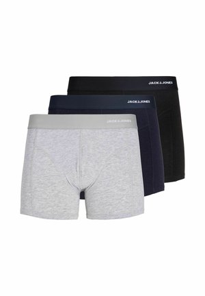 Jeune homme aux cheveux bouclés portant un boxer gris clair sur un fond clair uni.