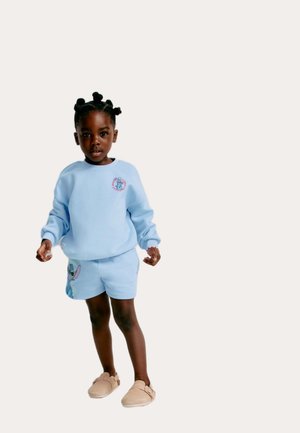 Helles blaues Sweatshirt und passende Shorts mit einem Charakterdruck. Weiches Baumwollmaterial, gerippte Bündchen und beige Slipper.