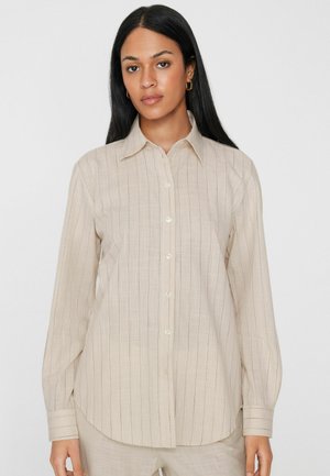 Frau mit langen dunklen Haaren, die goldene Creolen und ein beigefarbenes Hemd mit feinen vertikalen Streifen trägt, steht vor einem schlichten weißen Hintergrund.