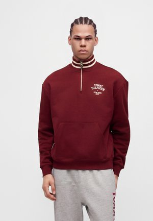 Млад мъж с плитки, облечен с бордова суитшърт на Tommy Hilfiger с цип до средата на гърдите и светлосиви спортни панталони, стои пред едноцветен фон.