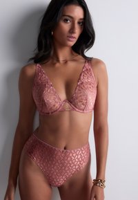 Femme portant un soutien-gorge en dentelle rose transparent et des culottes taille haute assorties, se tenant contre un mur blanc, avec des bracelets en or à son poignet.