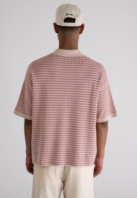 Short-sleeve beige knit shirt featuring a red triangular pattern, with a contrasting collar and wide sleeves. Worn with cream-colored pants.