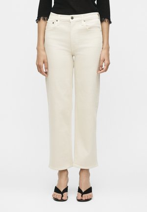 Γυναίκα που φοράει τζιν με ψηλή μέση και ίσια μπατζάκια σε εκρού απόχρωση, μαύρο τοπ και μαύρα ανοιχτά σανδάλια με τακούνι, στέκεται με χαλαρά χέρια.