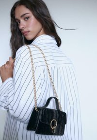 Borsa in velluto nero con tracolla in oro, caratterizzata da un design strutturato, manico superiore e un distintivo accento in metallo a forma di chiave.
