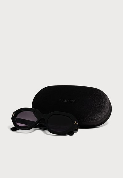 Lunettes de soleil ovales noires avec des verres foncés reposant devant un étui rigide noir de marque Tom Ford sur un fond clair.